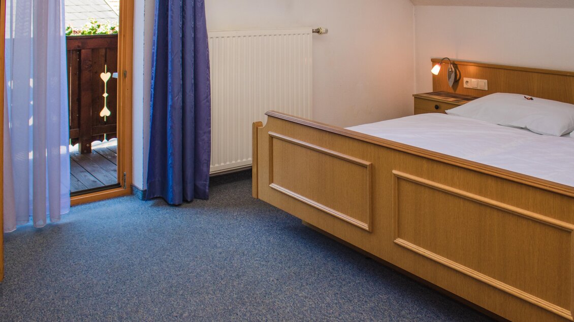 Ein gemütliches Schlafzimmer mit einem Holzbett und blauen Vorhängen. Der Zugang zu einem kleinen Balkon sorgt für Tageslicht und frische Luft. | © Gerhard Schernthaner