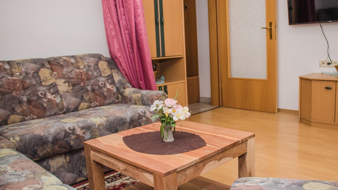 Ein gemütliches Wohnzimmer mit einem Holztisch und Sofas. Auf dem Tisch steht eine Blumenvasen und im Hintergrund befindet sich ein Fernseher. | © Gerhard Schernthaner