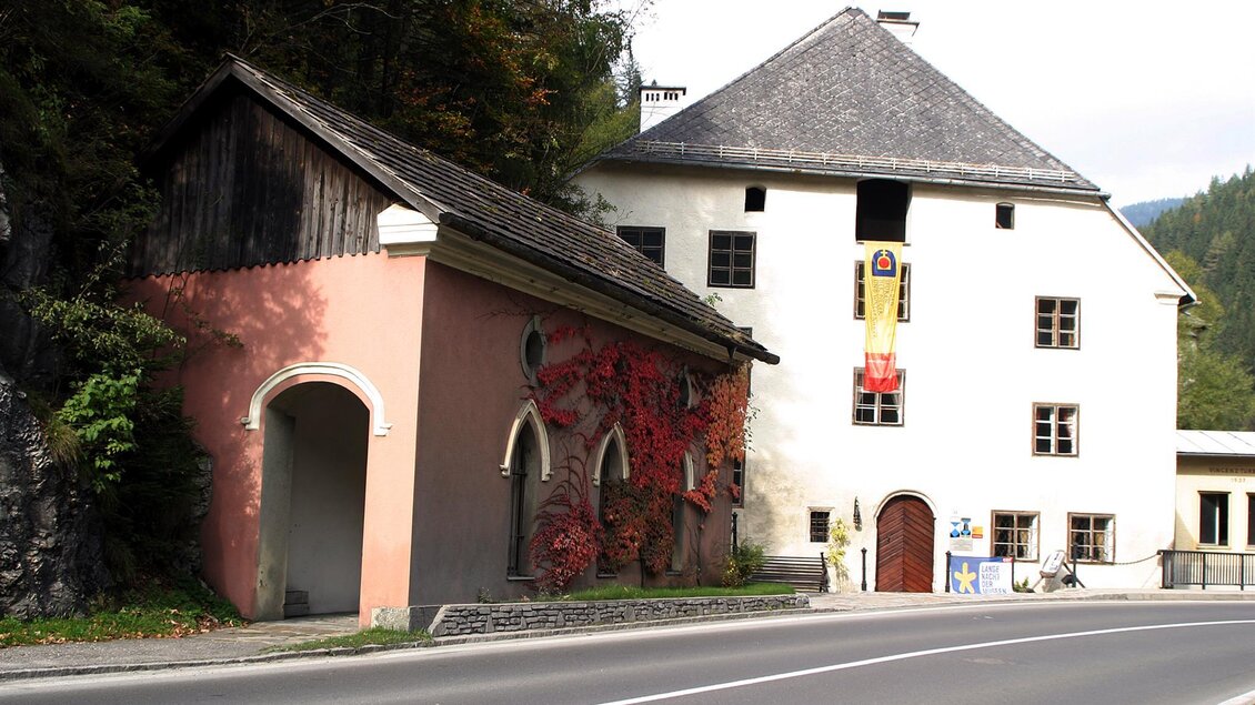 Eine historische Gebäudeansicht mit einem alten, verzierten Haus und einer kleineren, gemauerten Struktur. Die Umgebung ist von Bäumen und einer Straße umgeben. | © Marktgemeinde Thörl