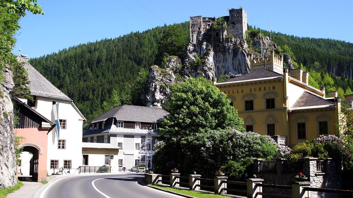 Eine malerische Straße führt an traditionellen Häusern vorbei, mit einem Blick auf eine beeindruckende Burg und umgebende Wälder. Die Landschaft ist grün und einladend. | © Marktgemeinde Thörl