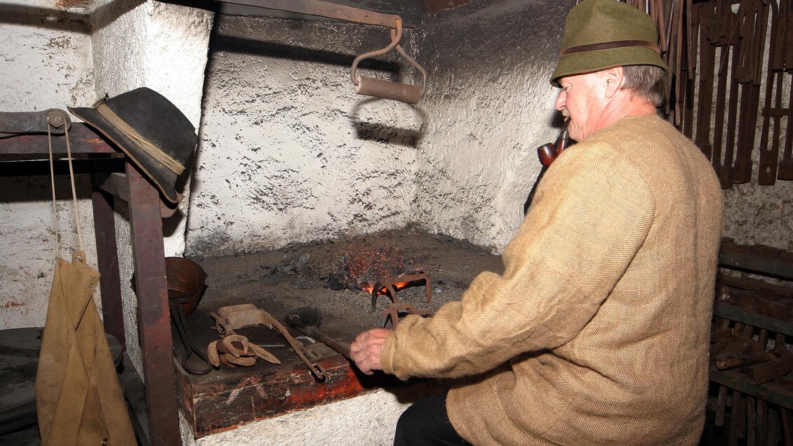 Ein Mann arbeitet an einem offenen Feuer in einer traditionellen Werkstatt. Er trägt einen Hut und ist mit einer handwerklichen Tätigkeit beschäftigt. | © Marktgemeinde Thörl