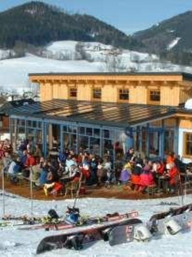A cozy mountain house in the mountains with many guests on the terrace. Surrounded by snow and ski equipment, the visitors enjoy the winter atmosphere. | © TV Hochsteiermark