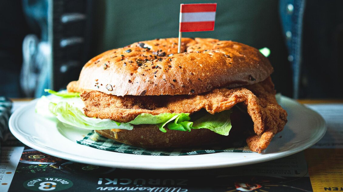 Ein knuspriger Burger mit einem Brötchen aus Samen und frischem Salat. Auf dem Burger steckt eine kleine Flagge in den Farben Rot und Weiß. | © 3 Goldene Kugeln