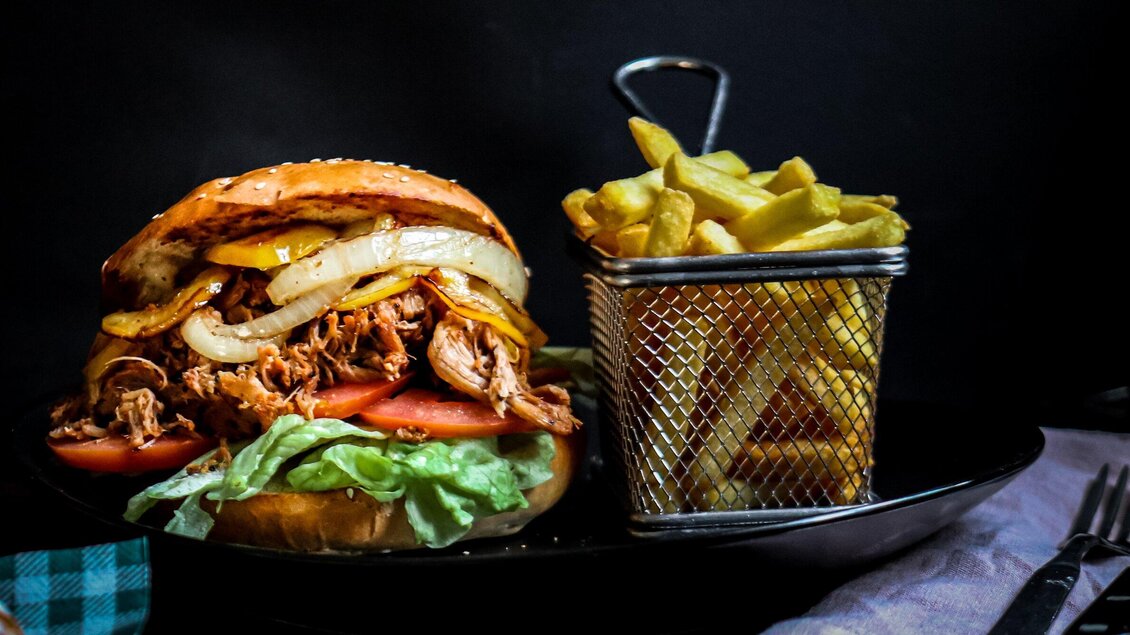 Ein saftiger Burger mit zartem Fleisch, Zwiebeln und frischem Gemüse. Dazu gibt es knusprige Pommes frites in einem kleinen Drahtkorb. | © 3 Goldene Kugeln