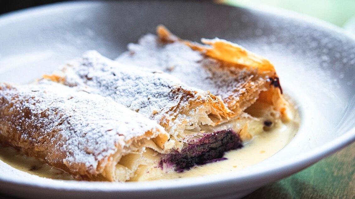 Ein Stück Apfelstrudel mit Puderzucker, serviert auf einer weißen Platte. Daneben ist eine Portion Vanillesoße. | © 3 Goldene Kugeln