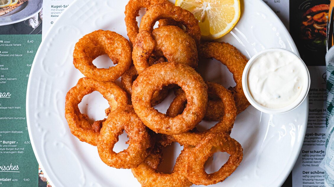 Ein Teller mit knusprigen Zwiebelringen, serviert mit einer Zitronenscheibe und einer Portion Dip. Die Zwiebelringe sind goldbraun und appetitlich angeordnet. | © 3 Goldene Kugeln