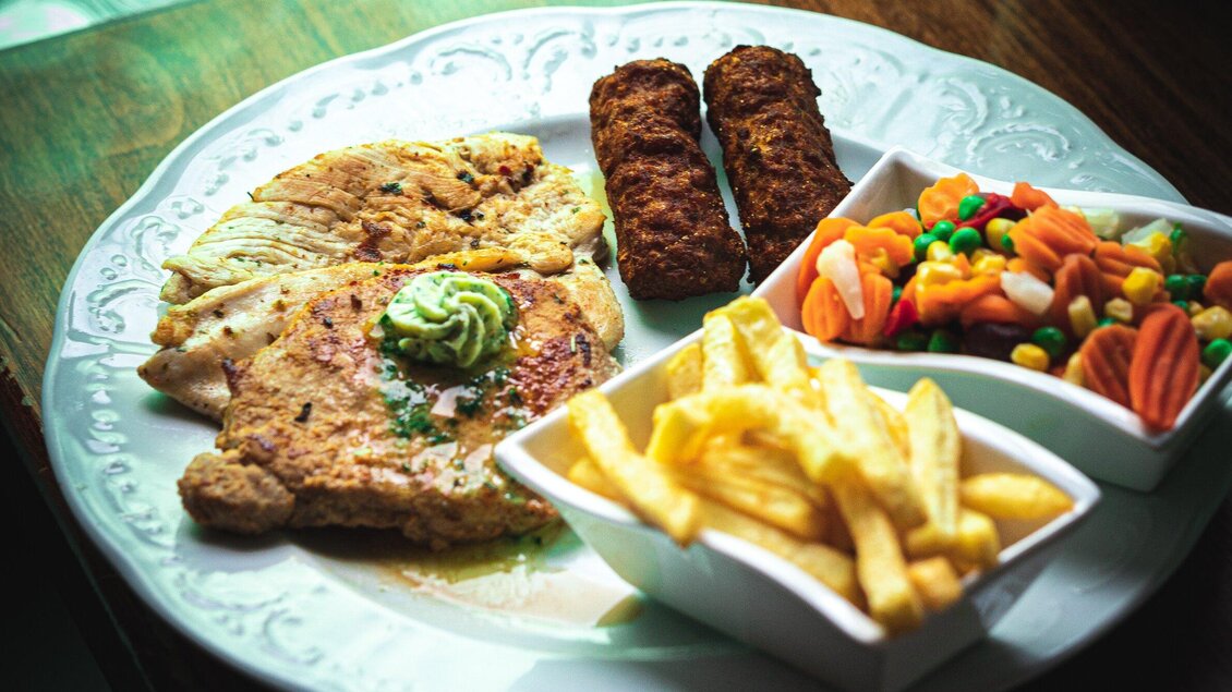 Ein Teller mit gebratenem Hühnchen, zwei Fischfrikadellen und Pommes frites. Dazu gibt es eine Schüssel mit buntem Gemüse. | © 3 Goldene Kugeln