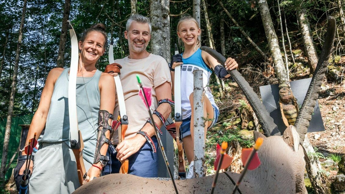 Eine Gruppe von drei Personen steht im Wald mit Bögen und Pfeilen. Sie lächeln und posieren vor einem Ziel. | © www.zloam.at
