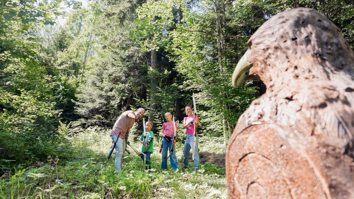 Eine Familie steht in einem Wald und betrachtet eine große Steinskulptur eines Adlers. Die Umgebung ist grün und sonnig, mit vielen Bäumen im Hintergrund. | © www.zloam.at