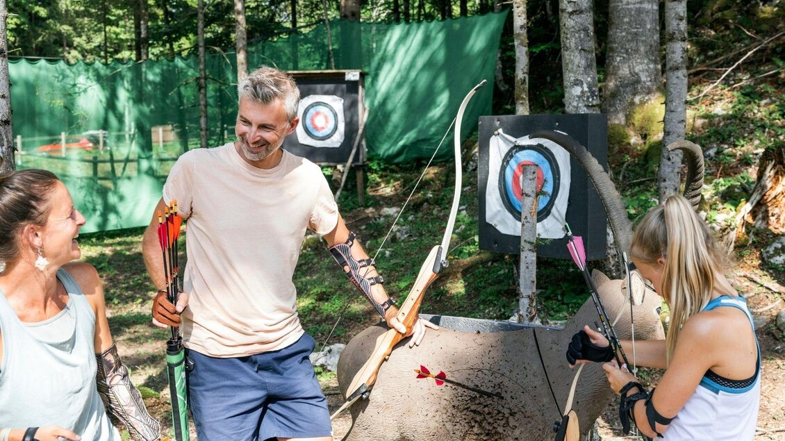 Drei Personen stehen beim Bogenschießen in einem Wald. Sie lachen und zeigen auf ihre Zielscheibe, während Pfeile in einem Ziel stecken. | © www.zloam.at