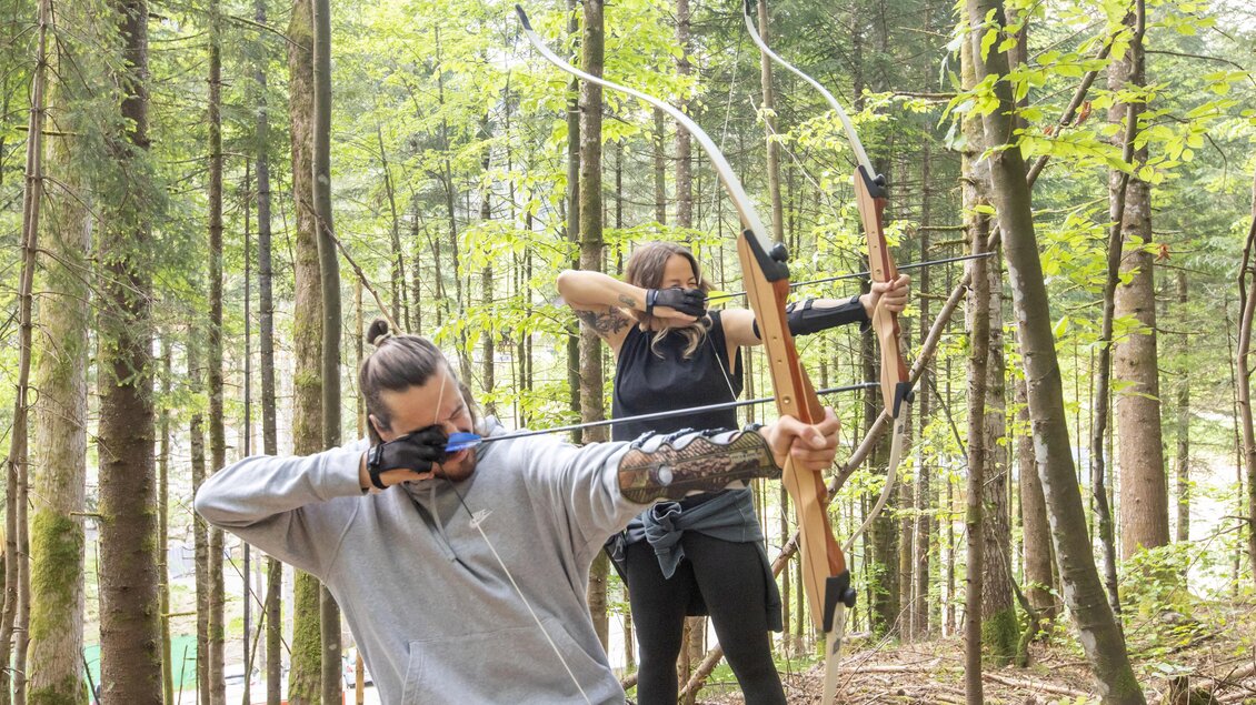 Zwei Personen schießen mit Bögen im Wald. Die Umgebung ist grün und von hohen Bäumen umgeben. | © Narzissendorf Zloam, www.zloam.at