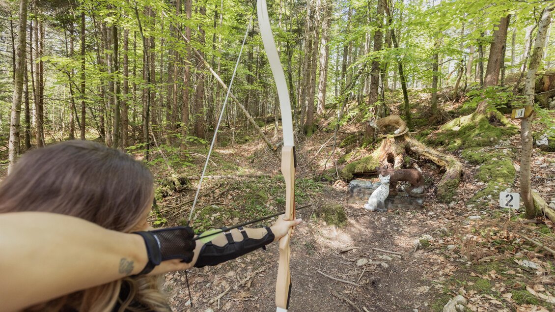 Eine Person zielt mit einem Bogen auf ein Ziel im Wald. Die Umgebung ist grün und natürlich, mit Bäumen und einem klaren Weg. | © Narzissendorf Zloam, www.zloam.at