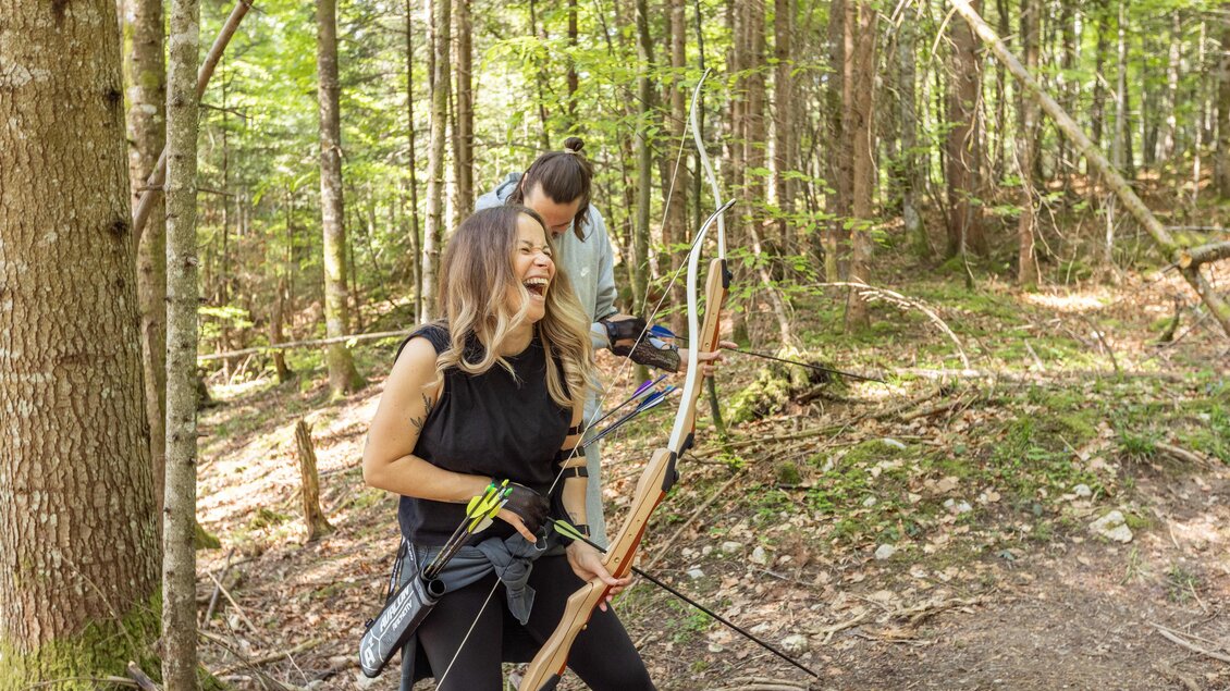 Eine Frau mit einem Bogen lächelt fröhlich im Wald. Im Hintergrund steht ein Mann, der ebenfalls einen Bogen hält. | © Narzissendorf Zloam, www.zloam.at