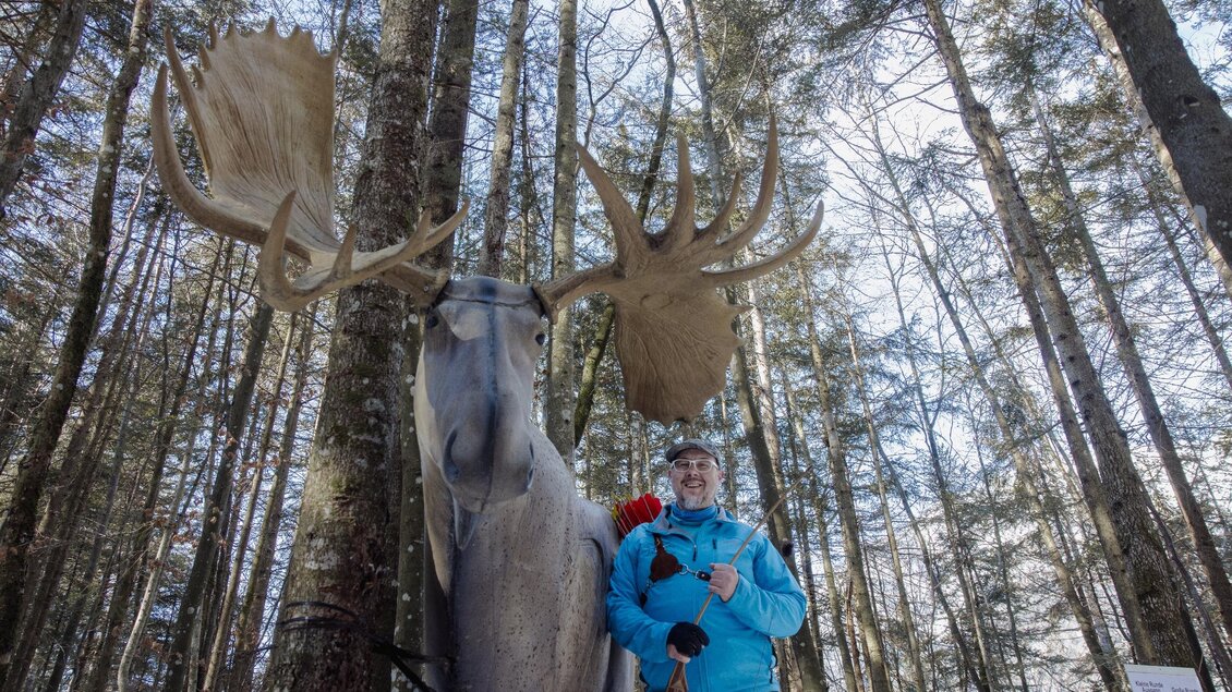 Ein Mann steht neben einer großen Elchstatue in einem Wald. Die Bäume um ihn herum sind hoch und verleihen der Szene eine natürliche Atmosphäre. | © Narzissendorf Zloam, www.zloam.at