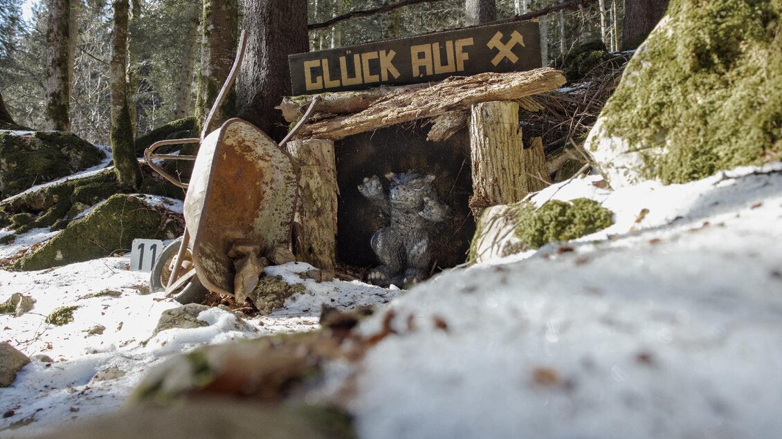 Ein kleiner Eingang zu einer Mine mit dem Schild „GLÜCK AUF“. Der Bereich ist von Bäumen und schneebedecktem Boden umgeben. | © Narzissendorf Zloam, www.zloam.at