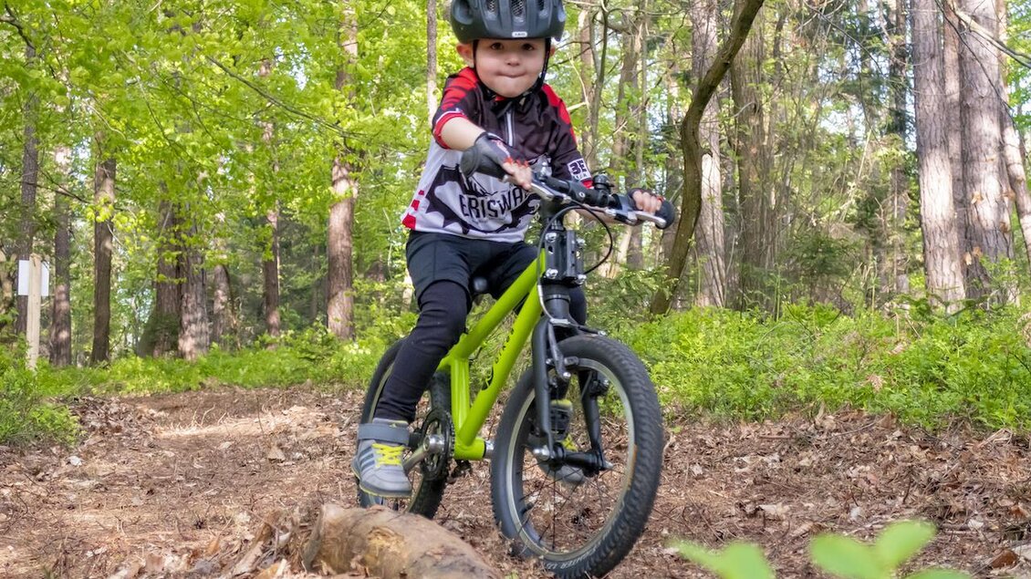 Ein Junge fährt mit einem grünen Fahrrad auf einem Waldweg. Im Hintergrund sind Bäume und grünes Laub zu sehen. | © 3Eiben Kids Bike Park