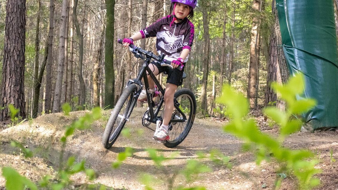 Ein Mädchen fährt mit ihrem Mountainbike auf einem Waldweg. Die Umgebung ist grün und sonnig, mit Bäumen im Hintergrund. | © 3Eiben Kids Bike Park