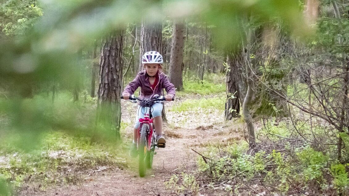 Ein Kind fährt mit dem Fahrrad auf einem schmalen Weg durch den Wald. Die Umgebung ist grün und von Bäumen umgeben. | © 3Eiben Kids Bike Park