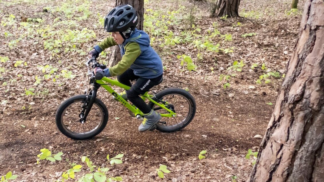 Ein Kind fährt mit einem grünen Fahrrad durch einen Wald. Der Junge trägt einen Helm und hat Spaß beim Radfahren auf dem Naturweg. | © 3Eiben Kids Bike Park
