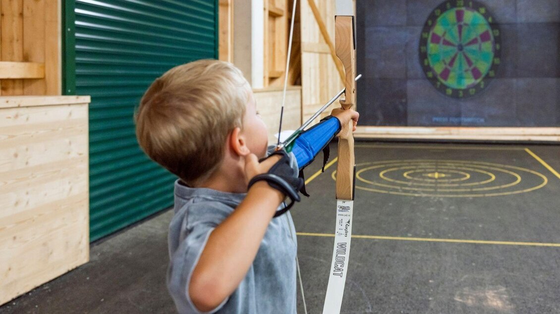 Ein Junge schießt mit einem Bogen auf ein digitales Ziel. Die Umgebung ist eine Trainingshalle mit Holzverkleidung. | © www.zloam.at