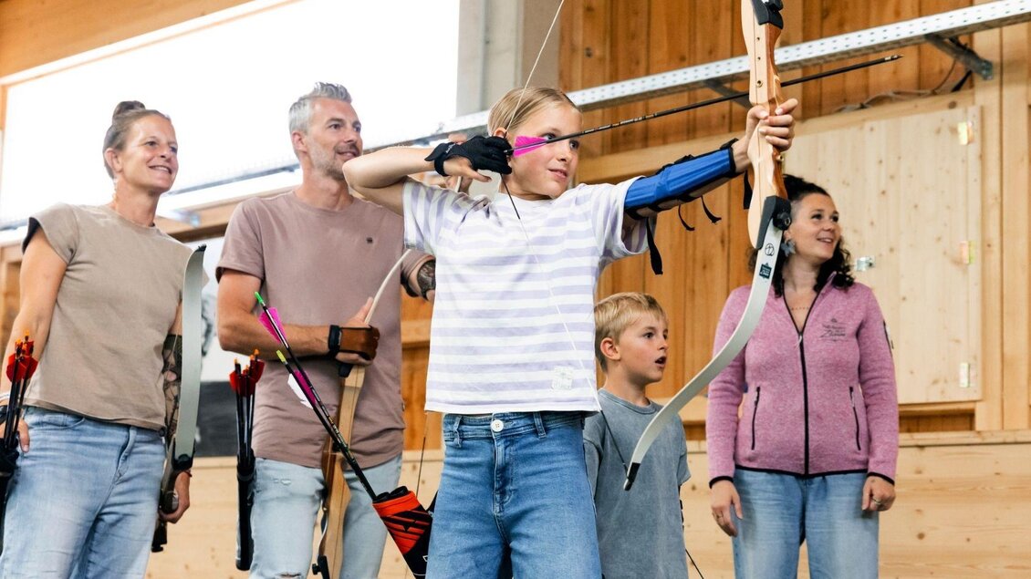 Eine Gruppe von vier Personen steht in einer Indoor-Archery-Anlage. Ein Junge zielt mit einem Bogen auf eine Zielscheibe. | © www.zloam.at