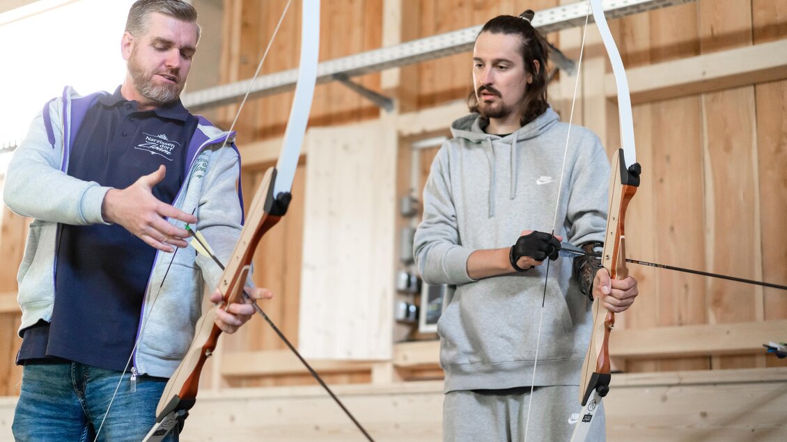 Zwei Männer stehen in einer Holzhütte und lernen Bogenschießen. Der Trainer zeigt dem Schüler, wie man den Bogen richtig hält. | © Narzissendorf Zloam, www.zloam.at