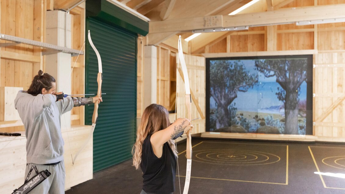 Zwei Personen üben den Bogenschießen in einem Holzraum. Im Hintergrund ist ein Projektorbild mit einer Landschaft zu sehen. | © Narzissendorf Zloam, www.zloam.at