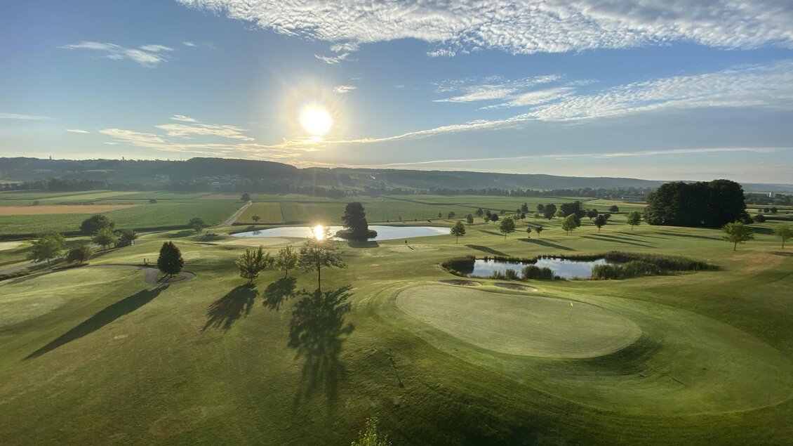 Ein schöner Golfplatz mit sanften Hügeln und Teichen. Die Sonne geht am Horizont auf und der Himmel ist teilweise bewölkt. | © Kindermann-Schön