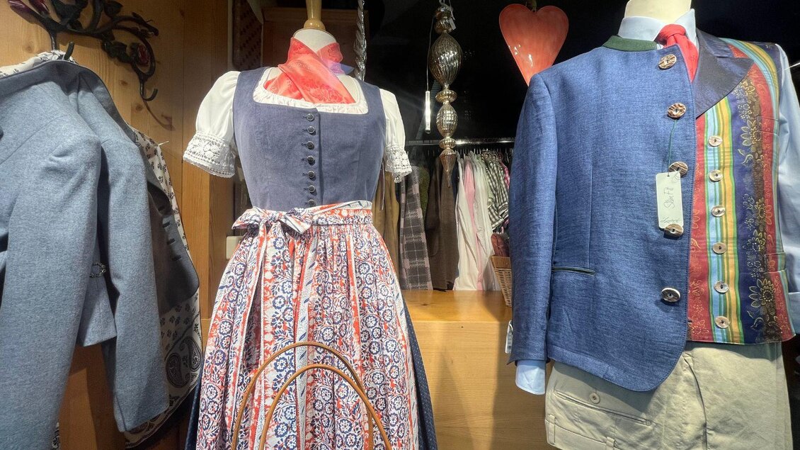 Eine traditionelle Tracht mit bunten Mustern und einer Schürze ist neben einem blauen Anzug ausgestellt. Im Hintergrund sind weitere Kleidungsstücke sichtbar. | © TVB Ausseerland-Salzkammergut_Nathalie Schröttenhamer