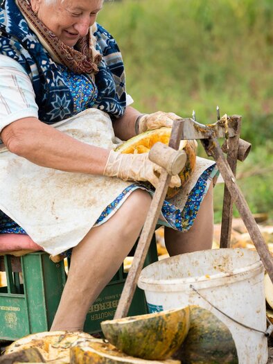 Eine ältere Frau schält Kürbisse auf einem Stuhl im Freien. Um sie herum liegen bereits einige bearbeitete Kürbisse und ein Eimer. | © Mias Photoart