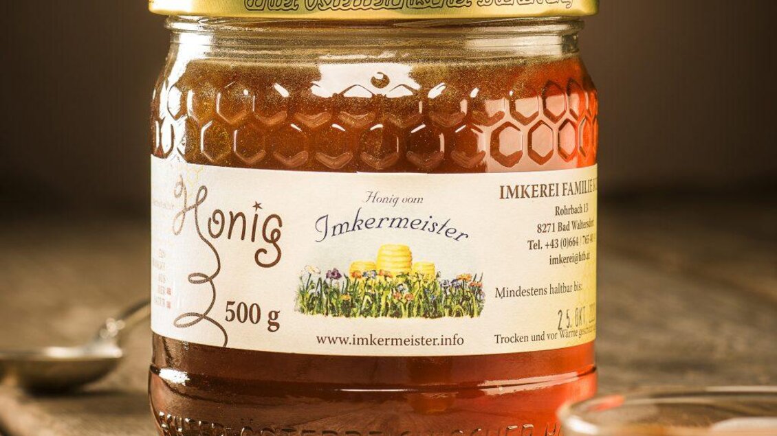 Ein Glas Honig mit einem goldenen Deckel auf einem Holzuntergrund. Die Etikette zeigt den Namen „Imkermeister“ und Informationen über das Produkt. | © Kurz