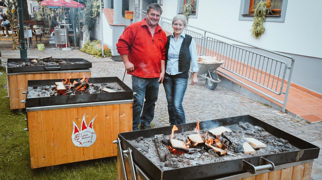 Ein Paar steht neben mehreren Grillstationen im Freien. Es raucht leicht und es gibt Holzfeuer in den Grills, um das Essen zuzubereiten. | © Pichler