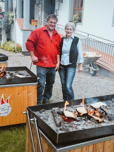 Ein Paar steht neben mehreren Grillstationen im Freien. Es raucht leicht und es gibt Holzfeuer in den Grills, um das Essen zuzubereiten. | © Pichler
