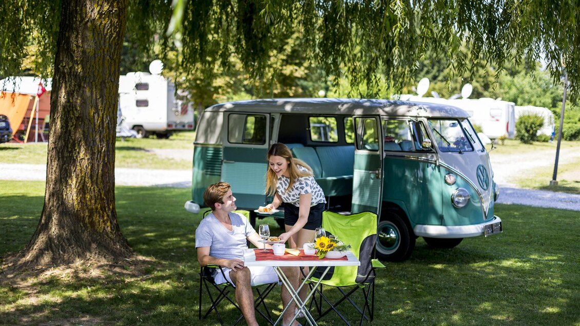Ein Campingplatz mit einem grünen VW-Bus im Hintergrund. Zwei Personen genießen ein Picknick unter einem Baum. | © Tomm Lamm