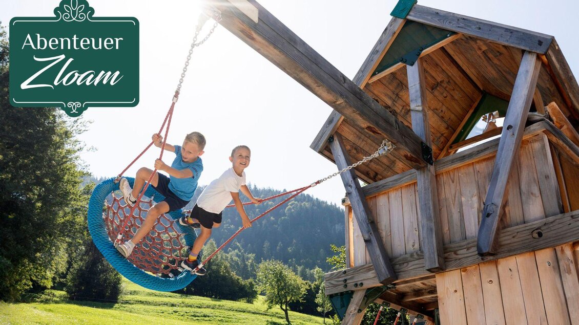 Zwei Kinder schwingen fröhlich auf einer großen Nestschaukel. Im Hintergrund sind eine bunte Natur und ein Spielplatz zu sehen. | © www.zloam.at