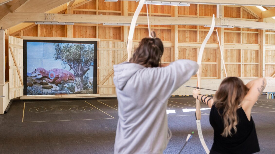 Zwei Personen üben Bogenschießen in einer Holzhalle. Im Hintergrund ist ein Bildschirm mit einem Bild von Wildtieren zu sehen. | © www.zloam.at