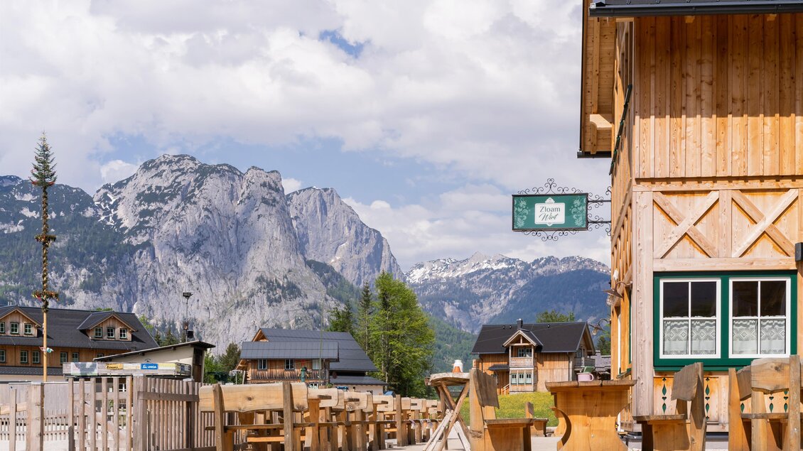 Ein traditionelles Holzhaus im Vordergrund mit einer schönen Berglandschaft im Hintergrund. Der Himmel ist teilweise bewölkt und bietet eine ruhige Atmosphäre. | © www.zloam.at