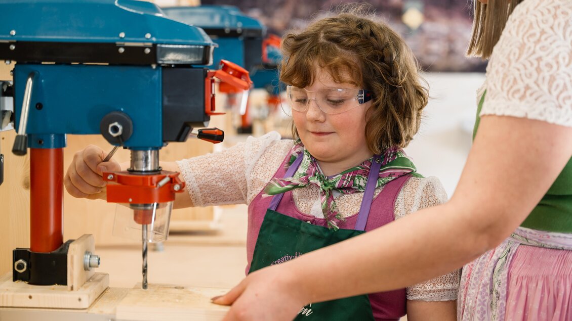 Ein Kind mit einer Schutzbrille bedient eine Bohrmaschine in einer Werkstatt. Eine weitere Person hilft ihm dabei, während sie Holz bearbeitet. | © Narzissendorf Zloam