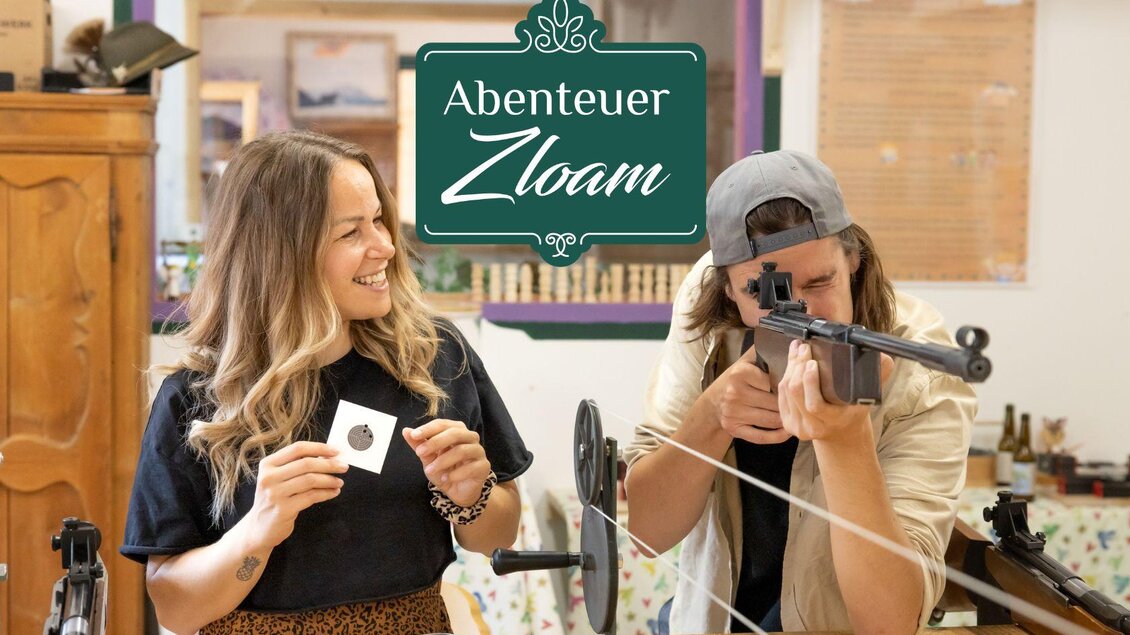 Ein Mann zielt mit einem Gewehr, während eine Frau lächelnd daneben steht und ein Objekt in der Hand hält. Hinter ihnen ist ein Schild mit dem Text "Abenteuer Zloam" sichtbar. | © www.zloam.at