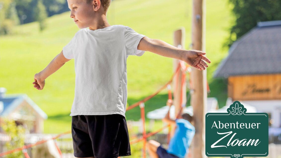 Ein Junge balanciert auf einer Slackline im Freien. Im Hintergrund sind grüne Wiesen und Spielgeräte zu sehen. | © www.zloam.at