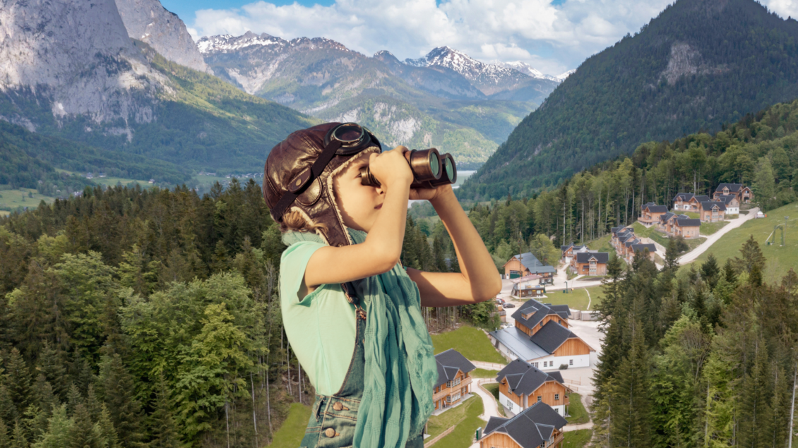 Ein Kind mit einer Fliegerbrille schaut mit einem Fernglas in die Ferne. Im Hintergrund sind Berge und eine kleine, malerische Siedlung zu sehen. | © www.zloam.at
