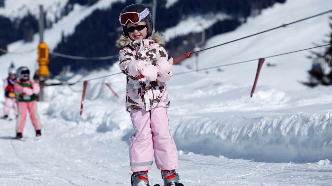 Ein kleines Mädchen fährt Skier auf einem verschneiten Hang. Im Hintergrund sind weitere Kinder und schneebedeckte Berge zu sehen. | © Narzissendorf Zloam www.zloam.at