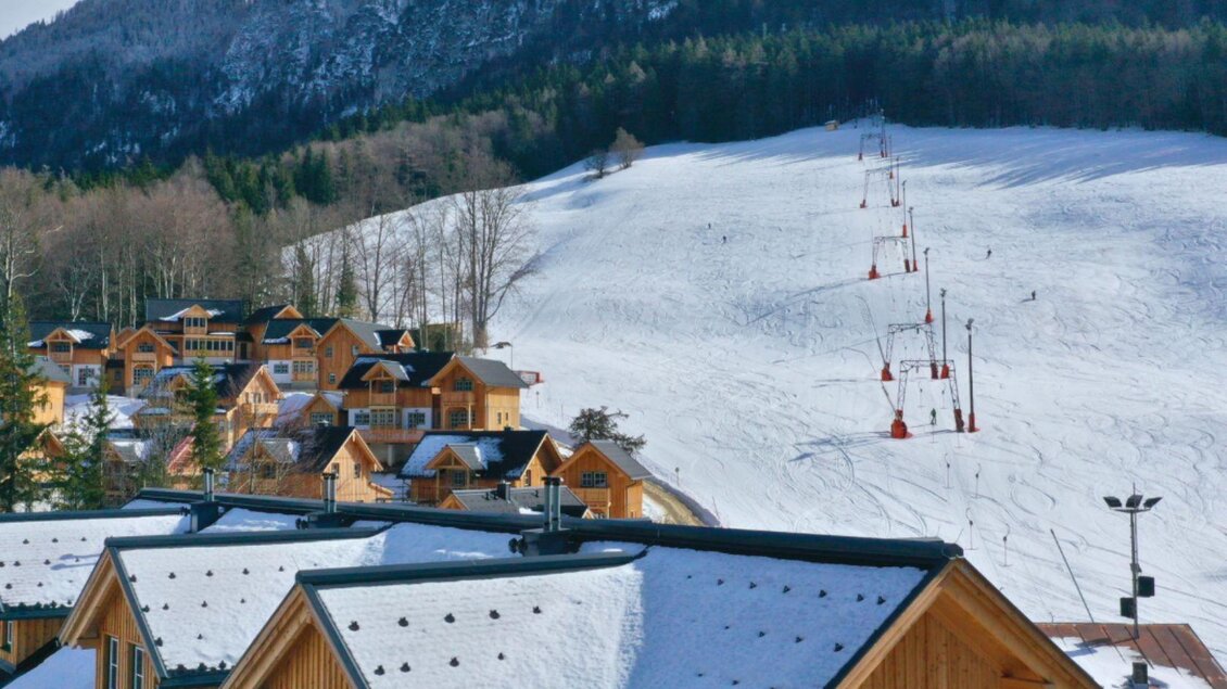 Eine verschneite Landschaft mit Skilift und umliegenden Hütten. Im Hintergrund sind bewaldete Berge sichtbar. | © Narzissendorf Zloam www.zloam.at