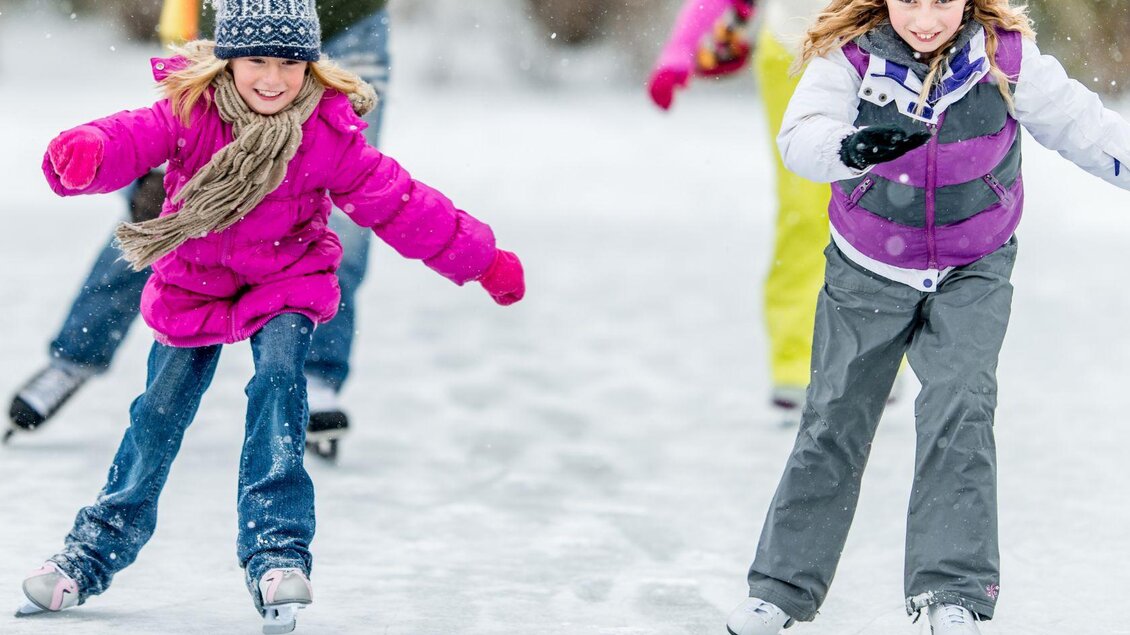 Zwei fröhliche Kinder in bunten Winterkleidungen gleiten auf einer Eislauffläche. Es schneit leicht und die Atmosphäre ist winterlich und festlich. | © Narzissendorf Zloam www.zloam.at
