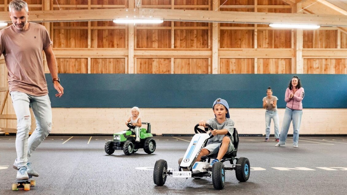 Eine fröhliche Szene mit Kindern, die in kleinen Go-Karts fahren, während Erwachsene zuschauen. Die Umgebung ist weitläufig und hell, mit Holzverkleidung an den Wänden. | © www.zloam.at