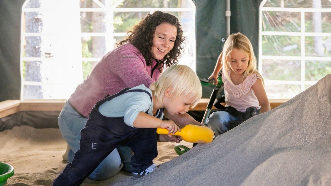 Eine Mutter spielt mit ihren zwei Kindern in einem Sandkasten. Die Kinder bauen mit Sandspielzeug, während sie fröhlich lachen. | © www.zloam.at