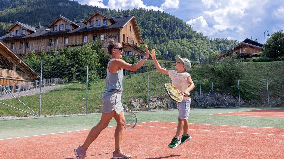 Eine Frau und ein Junge spielen Tennis auf einem Platz in den Bergen. Im Hintergrund sind Holzhäuser und die grüne Natur zu sehen. | © www.zloam.at
