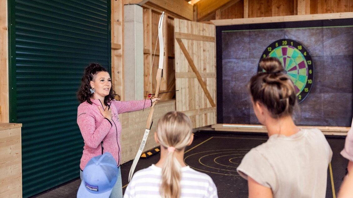 Eine Trainerin erklärt das Bogenschießen in einer gemütlichen Umgebung. Im Hintergrund ist eine Zielscheibe zu sehen, während die Teilnehmer aufmerksam zuhören. | © www.zloam.at
