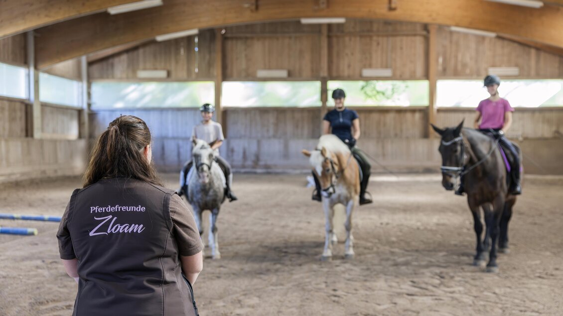Eine Reitstunde in einer Halle mit drei Reitern auf Pferden. Eine Trainerin beobachtet die Gruppe von hinten. | © www.zloam.at