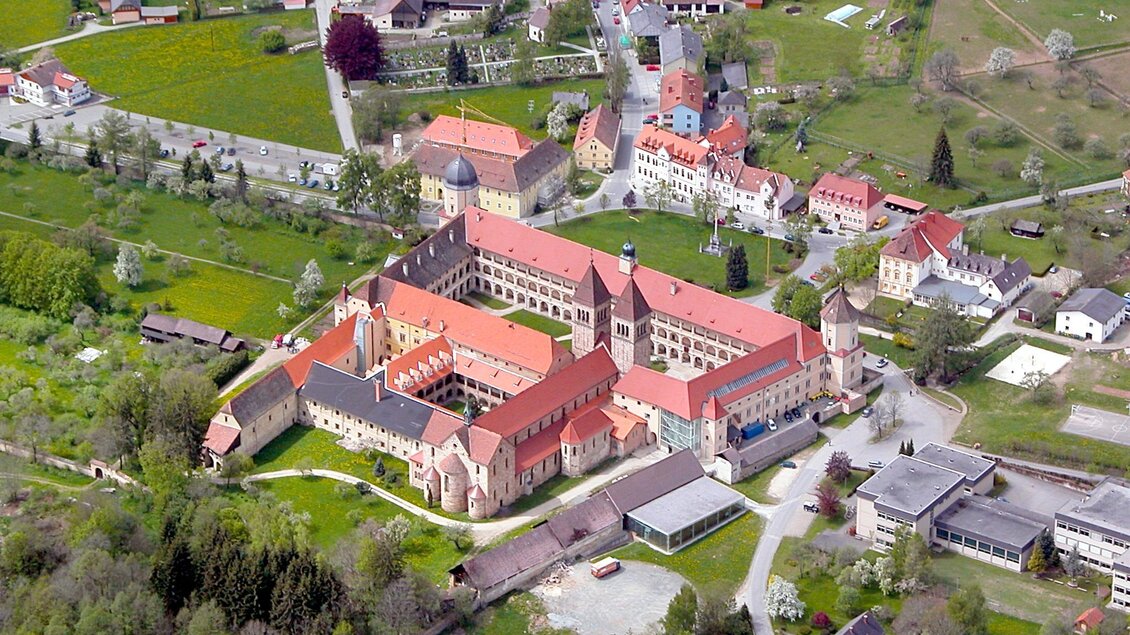 Eine beeindruckende Luftaufnahme eines großen Gebäudes mit rotem Dach umgeben von grüner Landschaft. Im Hintergrund sind kleine Dörfer und Felder zu sehen. | © Abtei Seckau